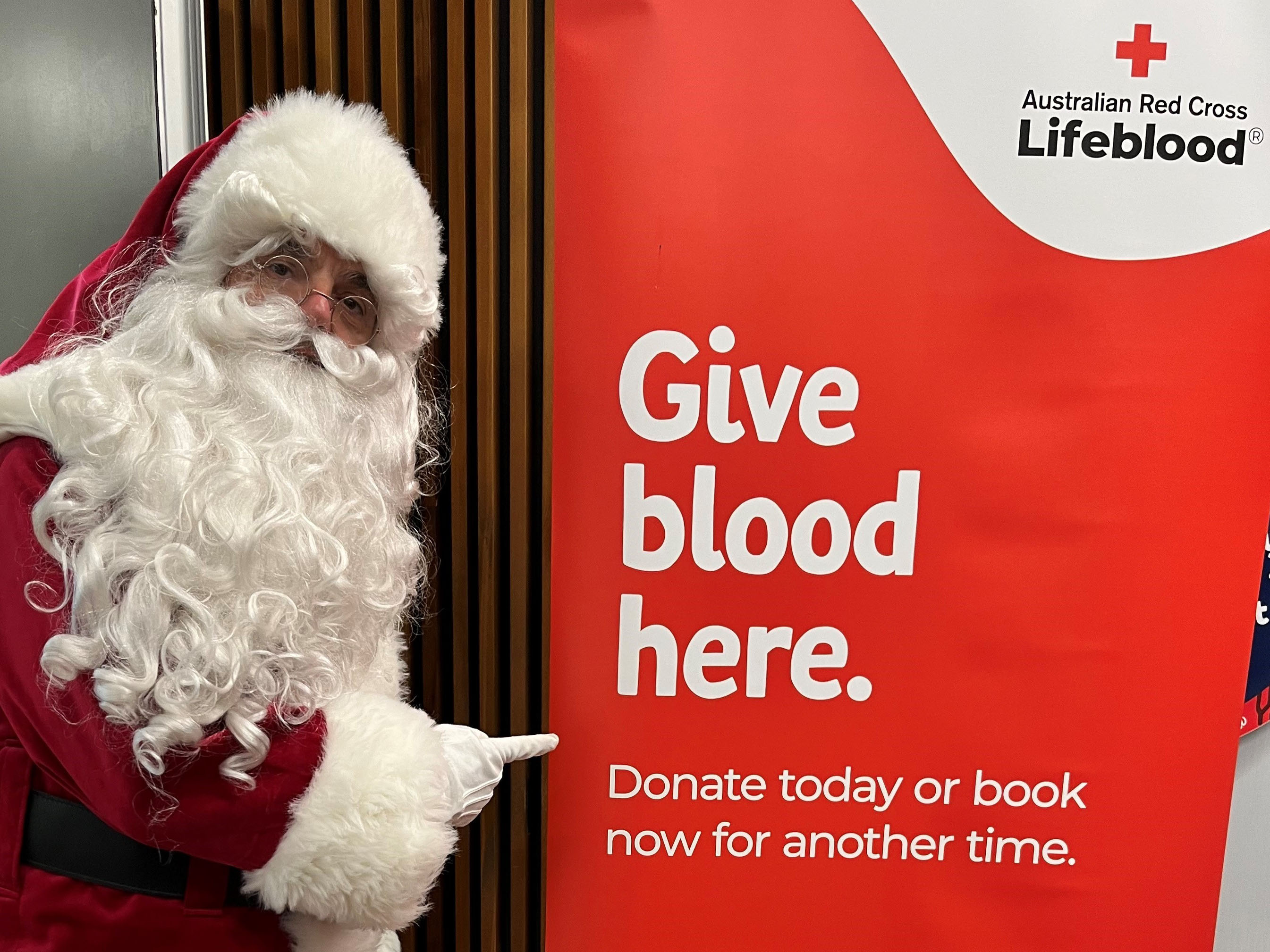 Man dressed as Santa in red suit and white beard pointing at sign which reads Give blood here 