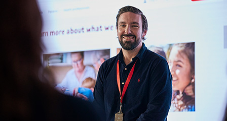 a staff member in smiling in front of a screen with a presentation on it