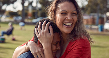 a mother is embracing her son and they are both laughing, a park can be seen in the background