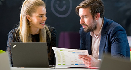two people talking at a desk with a laptop and printed pages