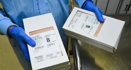 a lab worker is wearing a blue gown and gloves and is holding two boxes of blood packs marked B positive