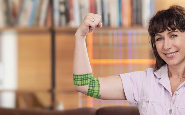 photo of Lisa Gorman at the camera holding up her right hand, she is wearing a light pink top and around her right elbow is the bandage she designed