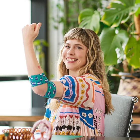 designer min pin is seated at a table wearing a colourful top and smiling, her left arm is in a flex pose and around her elbow is the bandage she designed