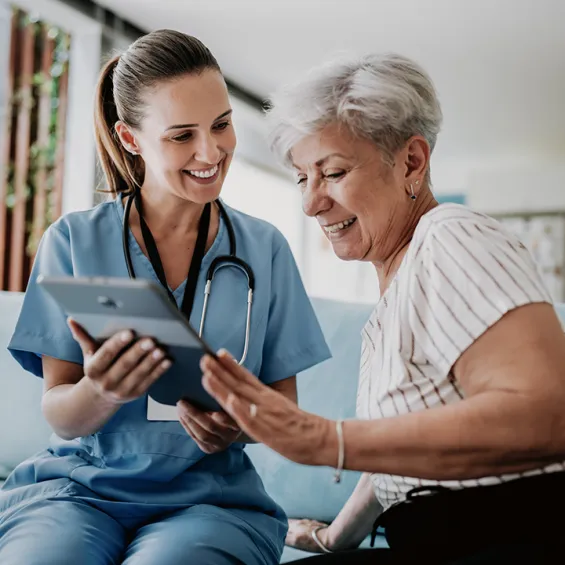 image of doctor in blue scrubs with stethoscope around neck sitting with elderly woman looking at an ipad