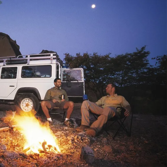 two men sitting and talking by a campfire with a four wheel drive parked behind them