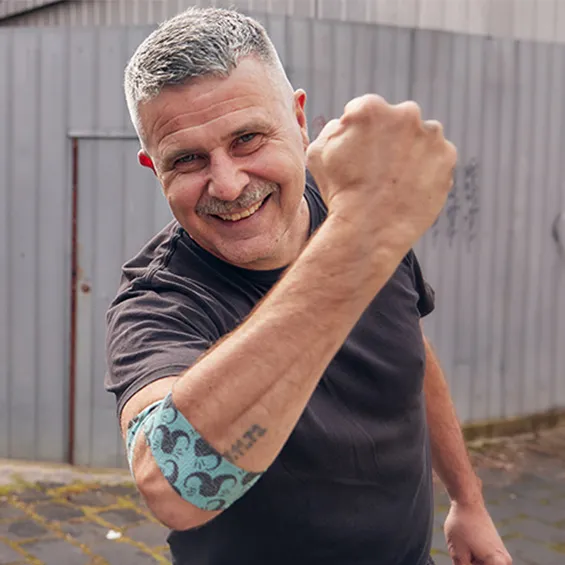 photo of Travis Garone smiling at the camera and holding up his right arm, around his elbow is the bandage he designed, he's wearing a black t-shirt and standing in a laneway