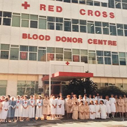 1980s colour photo of a large group of women, some of who are nurses, and a man in a tie standing outside a tall building saying "Red Cross Blood Donor Centre" 