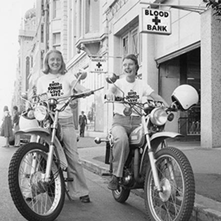 1970s black and white image. Two women on motorcycles wearing t-shirts saying "Blood Donors Love Life"
