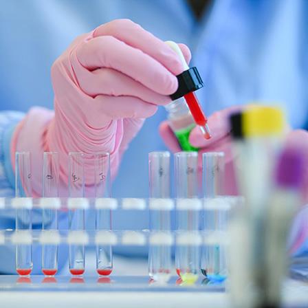 photo of a scientist wearing a blue gown and pink gloves holding a dropper over a row of test tubes