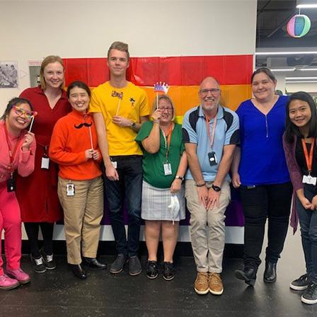 a group of people in an office standing in front of a pride flag and smiling at the camera