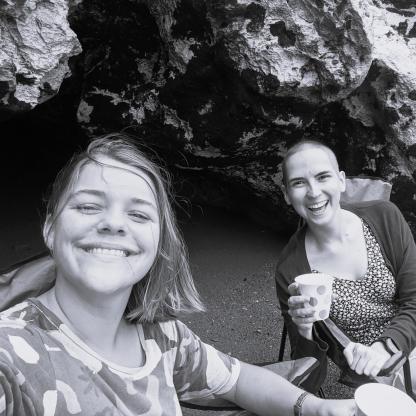 Two woman smiling happily at the camera. One had a shaved head and they are sitting on the beach under some rocks.