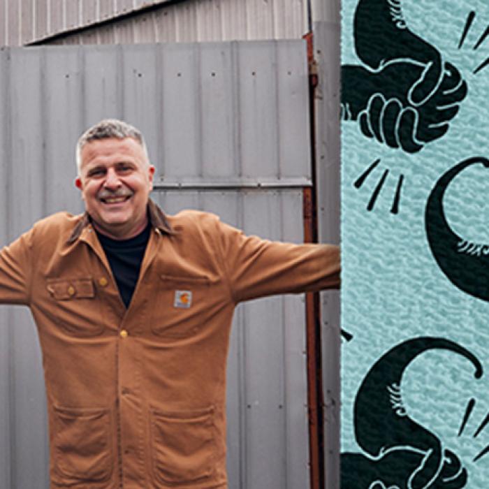 photo of Travis Garone smiling at the camera, he's wearing a brown jacket. The right hand side is an image of his design, it's on a light blue background and is of two hands in a stylised moustache shape